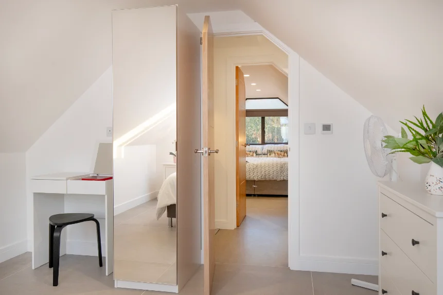 Mirrored wardrobe, small white dressing table and white chest of drawers in a bedroom under sloping eaves.
