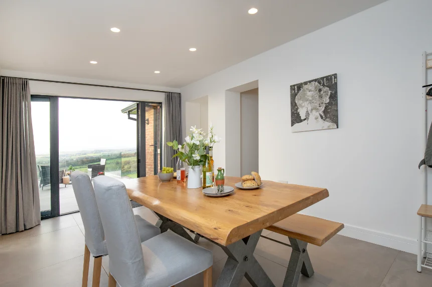Light wood dining table with chairs on one side and bench on the other in a bright modern room with full height windows opening on to patio