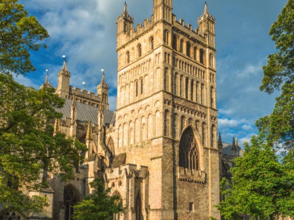 The main tower of Exeter Cathedral surrounded by trees against a blue sky