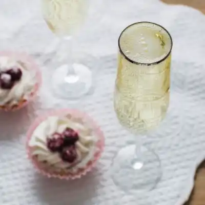 A glass of sparkling wine and two cupcakes with whipped cream and cherries on a white textured surface.